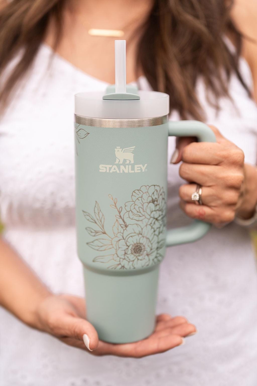 May include: A light green stainless steel travel mug with a floral design and a black handle. The mug has a clear lid with a straw and the words 'STANLEY' printed on the side.